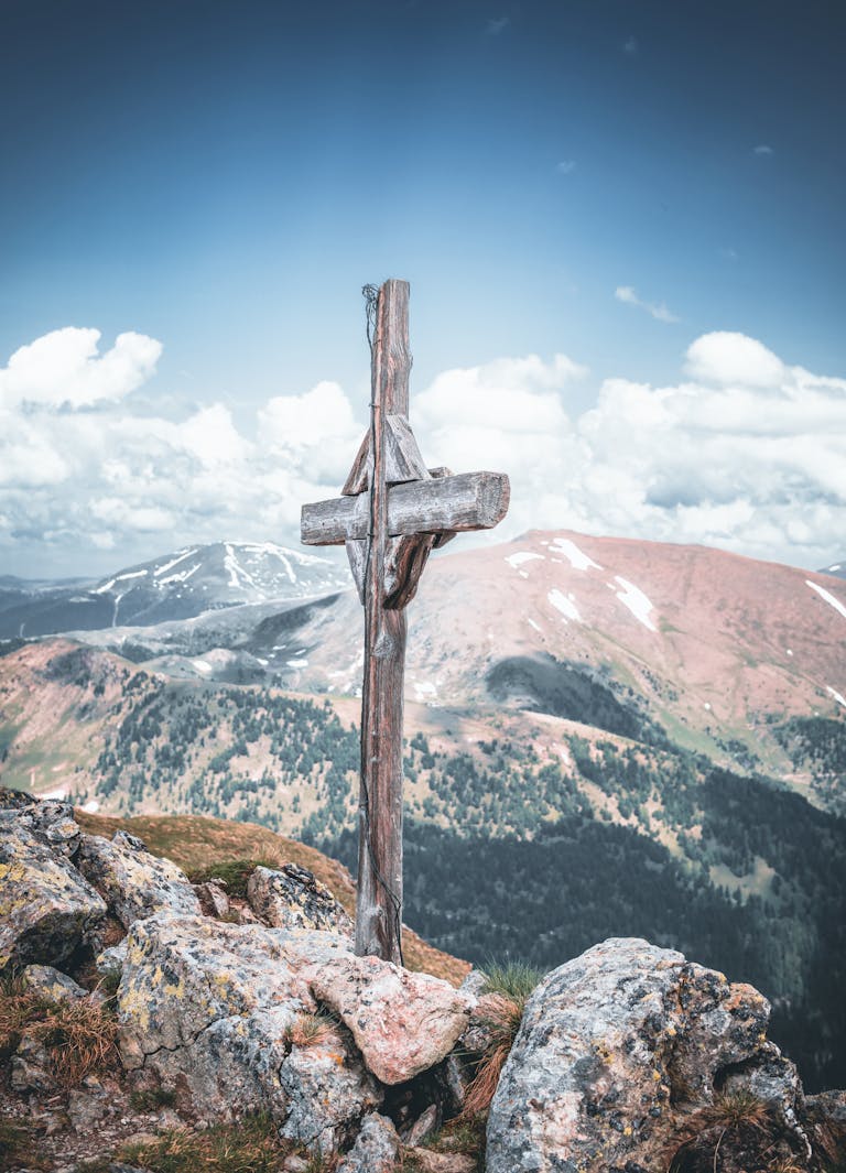 Rustic wooden cross on a rocky peak overlooking a scenic mountain range under a bright blue sky.